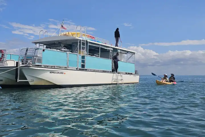 Boat with people onboard next to kayakers on calm ocean water under blue sky.