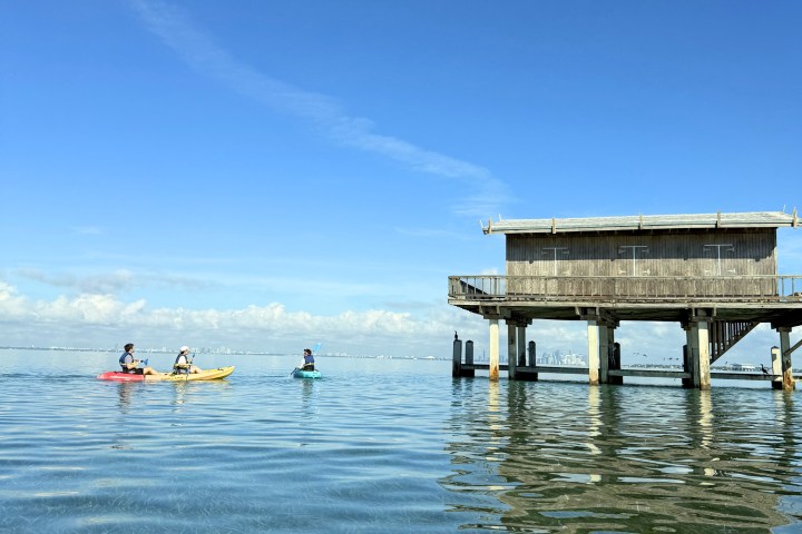 People kayaking near a stilt house on calm water under a clear blue sky.