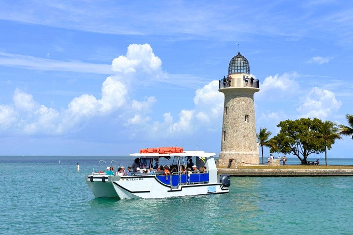 Boat near a lighthouse with people and palm trees on a sunny day.