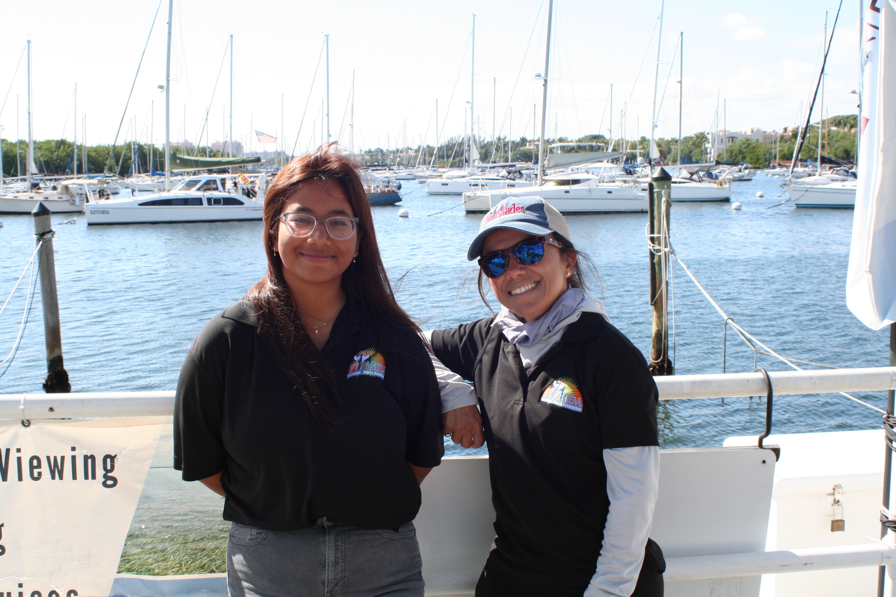 Two people smiling on a boat dock with sailboats in the background.