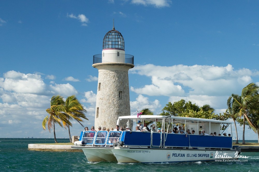 Boat with tourists near a lighthouse and palm trees on a sunny day.