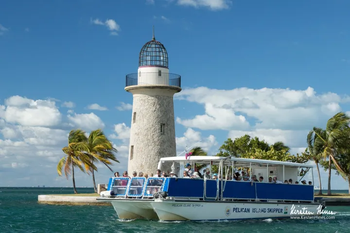 Boat with tourists near a lighthouse and palm trees on a sunny day.